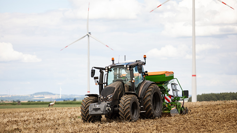 A Valtra G Series tractor pulls agricultural equipment across a harvested field. The scene is set in open countryside with multiple wind turbines in the background, highlighting a mix of farming and renewable energy. The sky is bright and slightly cloudy, giving the image a clean, natural atmosphere. A bird stands in the field near the machinery, adding a touch of wildlife to the rural landscape.